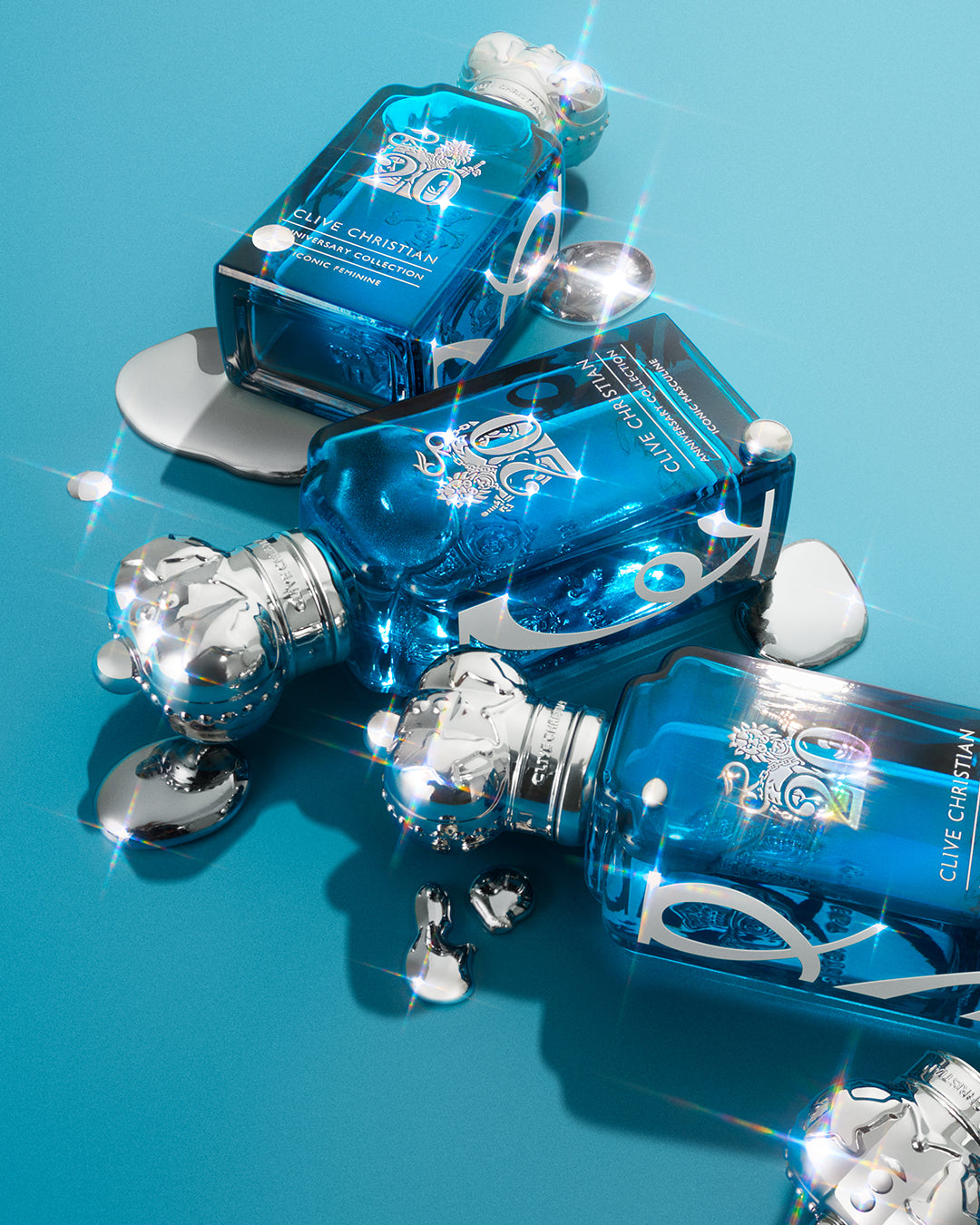 Three bottles of Clive Christian XX Anniversary Collection perfume in turquoise glass with silver crown caps lie on a turquoise surface with shimmering accents.