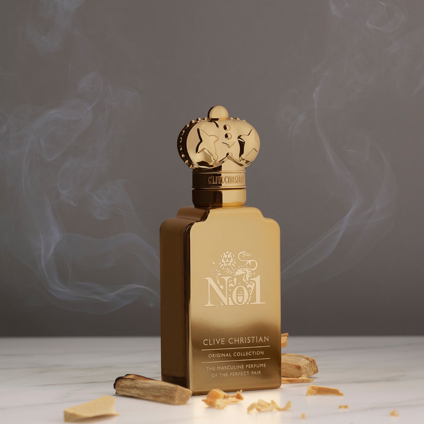 A gold bottle of Clive Christian No. 1 masculine perfume stands on a marble surface with aromatic wood chips, emitting a wisp of fragrant smoke.